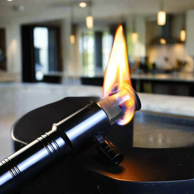 Black premium torch lighter emitting a strong precision flame, set in a modern kitchen background.